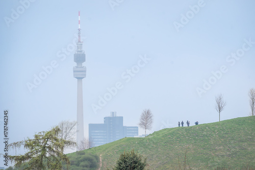 Wallpaper Mural Urban Dortmund landscape with communication tower and silhouettes of people on grassy hill under soft blue sky. Torontodigital.ca