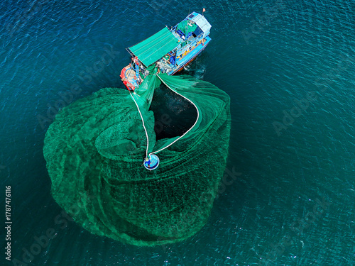 Aerial view of a fishing boat casting its emerald net into the deep blue sea, its circular form contrasting with the vessel's rigid structure, An Ninh Dong, Phu Yen, Vietnam.