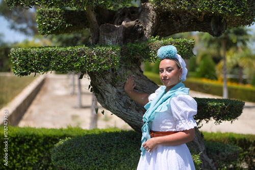 Carta da parati Young woman, beautiful, blonde, with typical Andalusian flamenco costume to go on pilgrimage in white with light blue shawl and flower, leaning on a tree trunk