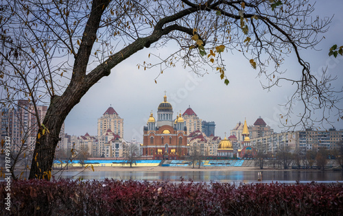 Cloudy evening view of Obolon Embankment in Kyiv with the Church of the Intercession and modern buildings reflected in the calm Dnipro River, framed by autumn branches and soft light.