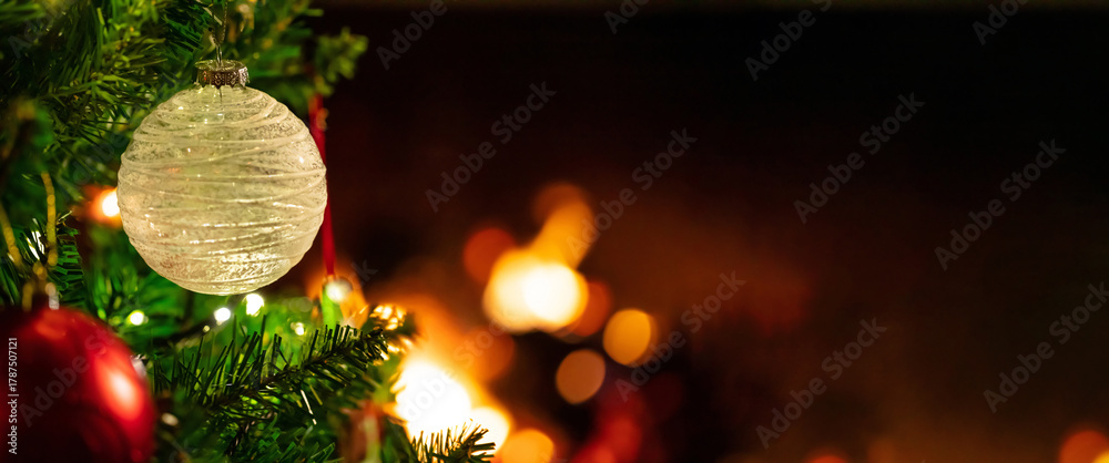 Naklejka premium Shimmering Christmas ornament hanging upon the green tree branch near a cozy fireplace glow, creating a festive and warm holiday mood