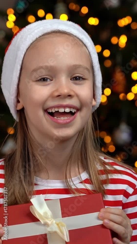 Portrait of happy girl in red santa hat looks at camera and laughs holding gift box and on background of Christmas tree. New Years present. Vertical shot