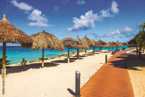 Fototapeta Naklejka Na Ścianę i Meble -  Beautiful view of wooden walkway and thatched palm leaf umbrellas on hotel beach located on Eagle Beach Caribbean Sea. Aruba.