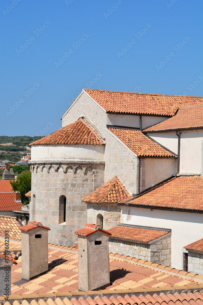 Fototapeta premium A detailed view of the terracotta tile rooftops and stone structure of the Cathedral of the Assumption of the Blessed Virgin Mary in Krk under a clear blue sky