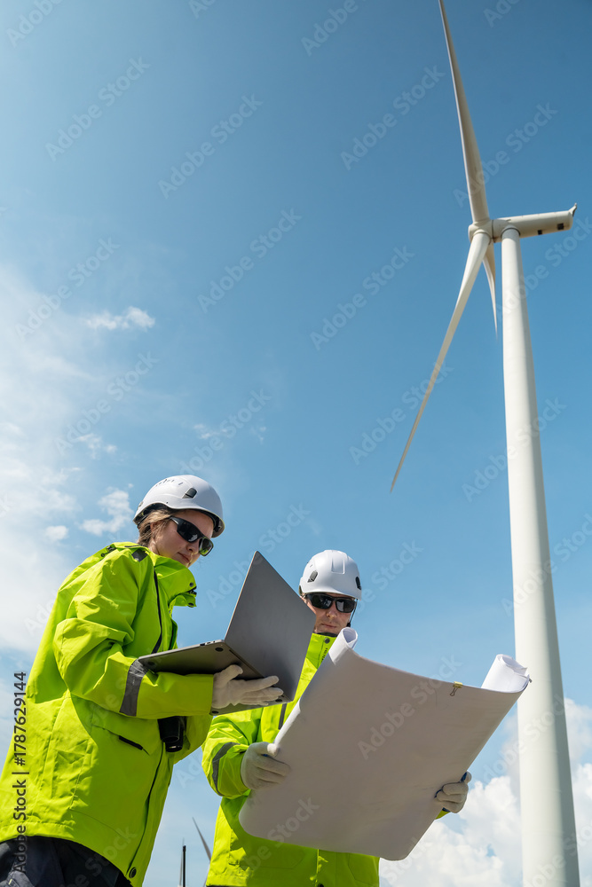 Fototapeta premium Two people are standing next to a wind turbine. One of them is holding a laptop. Low angle view of two technicians (man and woman) inspecting wind turbine structure.