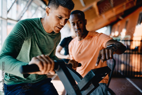 Trainer offering encouragement to a fit young man riding a stationary bicycle during a workout session at the gym