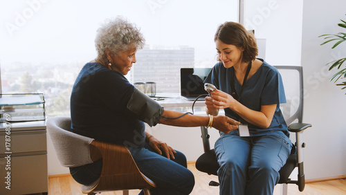 Nurse Wearing Scrubs In Hospital Office Checking Senior Female Patients Blood Pressure