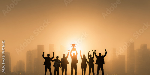 Silhouettes of business people celebrating success and teamwork against a city nightscape with one person holding a trophy high symbolizing achievement and unity.