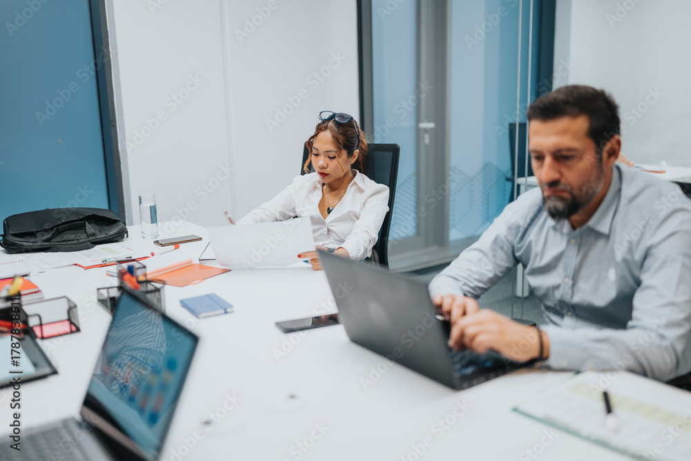 Fototapeta premium A modern office scene shows a woman reviewing papers while a man works on a laptop. Shared desk, notes, and devices convey focus, teamwork, and productive collaboration.
