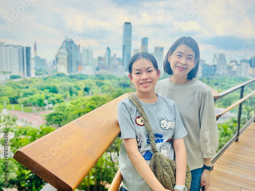 Asian preteen girl and middle-aged mother stand on a rooftop garden in Bangkok, smiling. The city skyline with tall buildings and lush greenery is visible in the background, under a partly cloudy sky