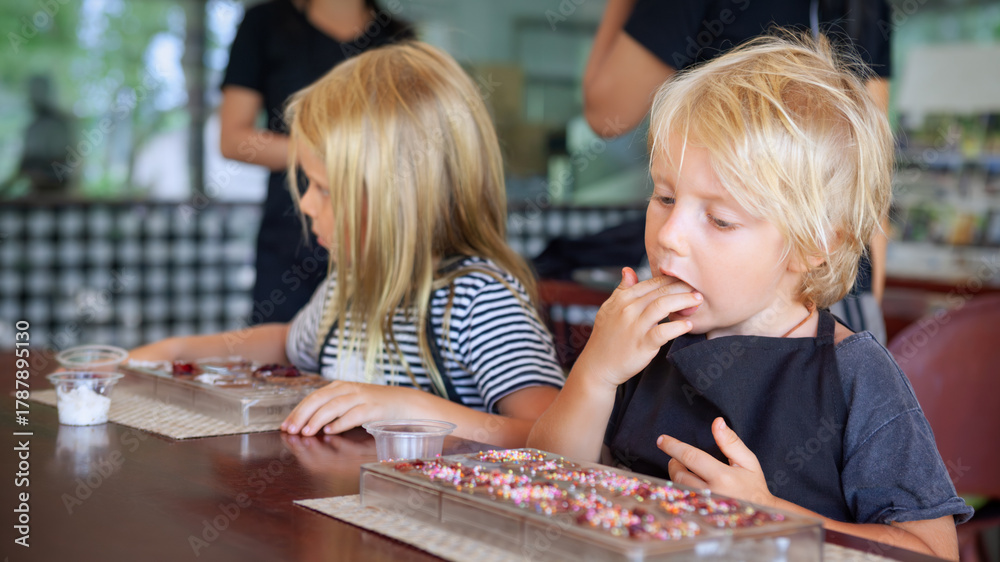 Fototapeta premium Two kids enjoy chocolate making class in Bali. Little boy happily tastes his sweet creation while girl focuses on decorating. A fun and flavorful family experience during travel