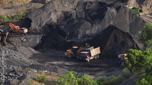 Large open-pit quarry with massive excavators and dump trucks loading and hauling coal and ore, showing heavy machinery, earthmoving operations and dusty industrial landscape