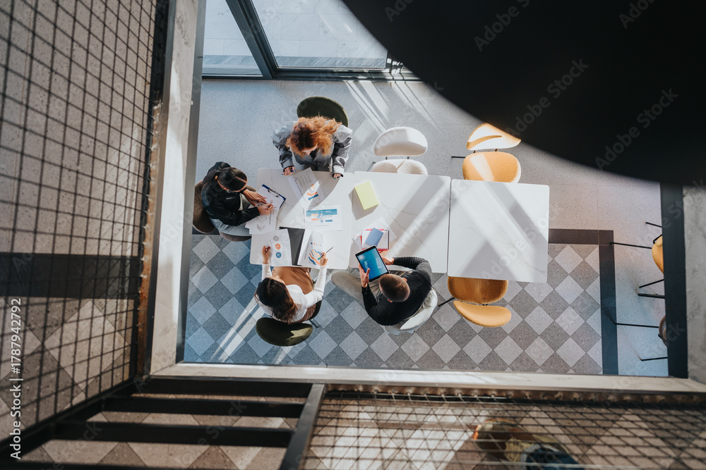 Obraz premium A group of professionals gathers around a table to review charts, documents, and digital devices during a collaborative business meeting in a bright, modern office.