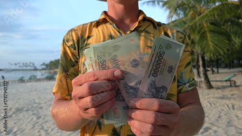 Man counting Vietnamese dong banknotes on tropical beach under palm trees in Vietnam. Perfect for travel, finance, and lifestyle concepts about wealth and freedom in Asia.