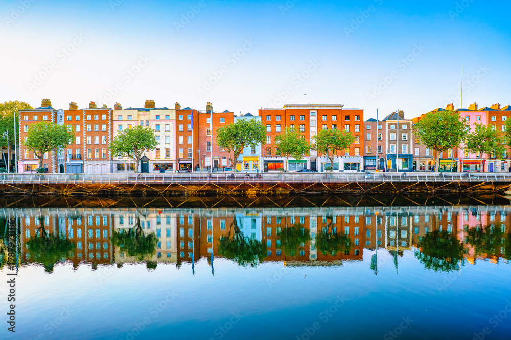 Fototapeta premium A colorful row of historic buildings along the River Liffey in Dublin is beautifully reflected in the calm water under a clear blue sky.