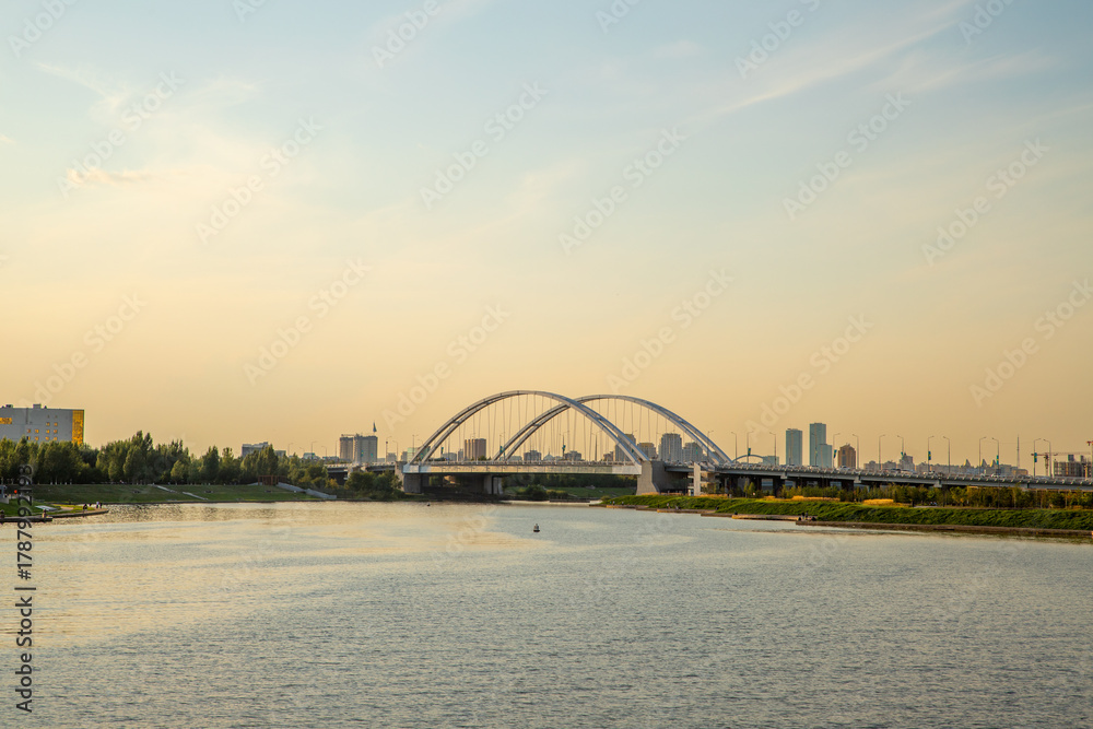 Naklejka premium Sunset view over a river with a modern steel arch bridge and city skyline in the background. Astana, Kazakhstan, Argar Bridge over the Ishim River