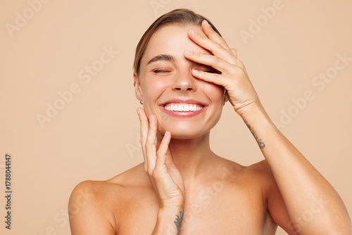 Close up fun beautiful half naked topless young woman 20s with nude make up close eyes touch face isolated on beige color background studio portrait. Skin care healthcare cosmetic procedures concept.