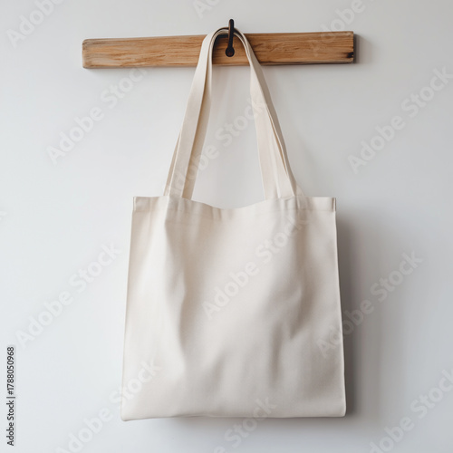 Minimalist off-white fabric tote bag hanging on wooden hook against white wall, mockup