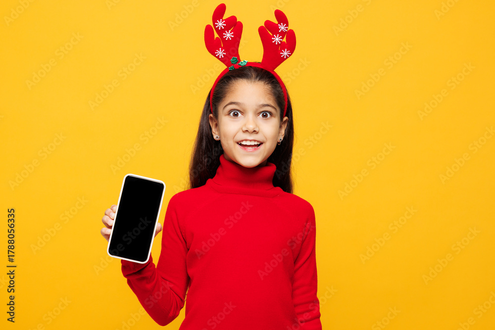 Naklejka premium Merry little girl wear red clothes fun decorative deer horns on head posing hold use blank screen mobile cell phone isolated on plain yellow background. Happy New Year Christmas celebration concept.