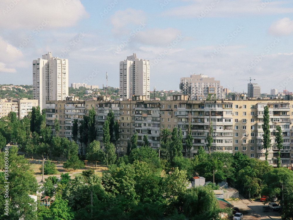 Obraz premium Cityscape of residential buildings with trees under a cloudy sky. This image shows a view of a city with apartment buildings and lush green trees on a sunny day