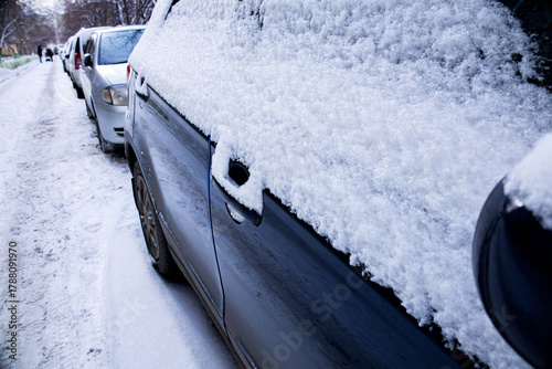 Fototapeta Naklejka Na Ścianę i Meble -  Snow covers parked cars along a quiet street in winter, creating a peaceful and serene atmosphere in a small town