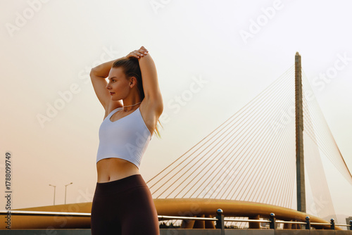 Young caucasian female stretching in front of modern bridge at sunrise