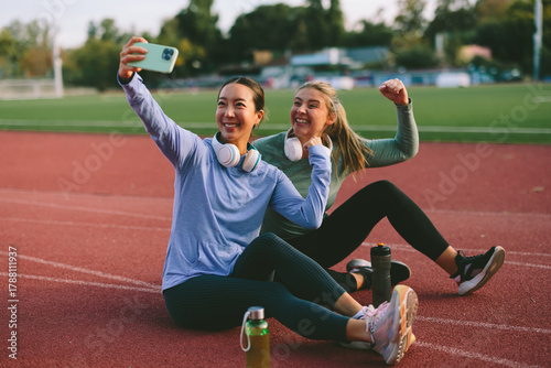 Diverse female friends and athletes, one Asian and one Caucasian, take a joyful selfie on a running track with one friend flexing her fist in a sign of fitness triumph or success
