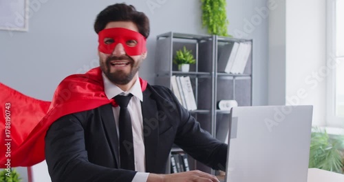Cheerful positive man dressed in superhero costume with mask and cape, showing biceps, demonstrating that he is powerful, bragging about his achievements, working on laptop computer in office room