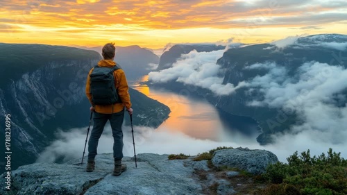 A hiker gazes over a fjord at dawn, clouds nestle amidst steep mountains, golden sky above