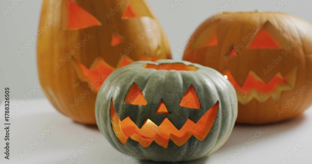 Naklejka premium Glowing green-gray carved pumpkin sitting on white tabletop in studio, with two lit orange pumpkins