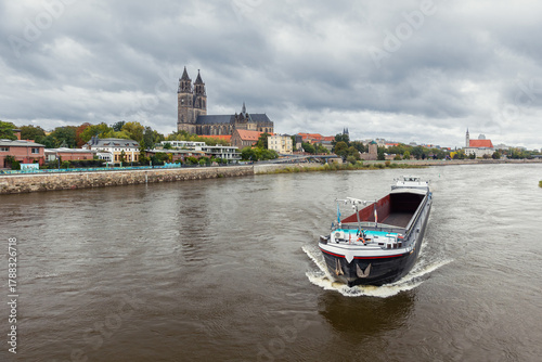 Cargo barge ship sails river Elbe historic Magdeburg city skyline cathedral Dom towers and colorful autumn trees. Overcast sky calm water create tranquil atmosphere European river inland transport