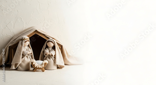 Minimalist Nativity scene with serene, faceless figures of Mary and Joseph in soft beige robes, standing beside a wooden manger under a draped cloth. Set against a textured off-white wall.