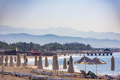 Fototapeta Naklejka Na Ścianę i Meble -  Sunbeds with towels and white umbrellas along morning shoreline, curved beach with swimmers and palm trees against hazy mountain backdrop in backlight. Sorgun, Antalya, Turkey.

