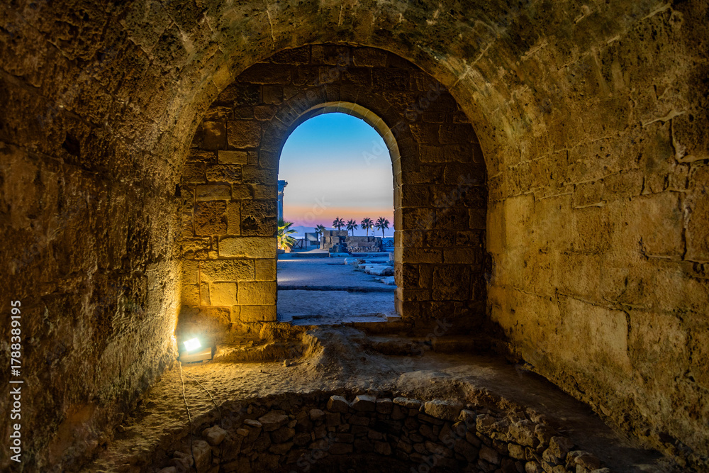 Obraz premium Byzantine stone vault with archway framing view of Apollo Temple and palm trees against orange sunset sky at ancient archaeological site. Side, Antalya, Turkey.