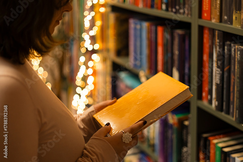 Woman holding new wrapped book in cozy bookstore with holiday bokeh lights.
