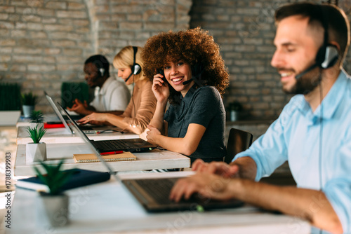 Smiling female customer service agent or call center employee sits at her desk with a headset, working enthusiastically in a modern, diverse office with her colleagues