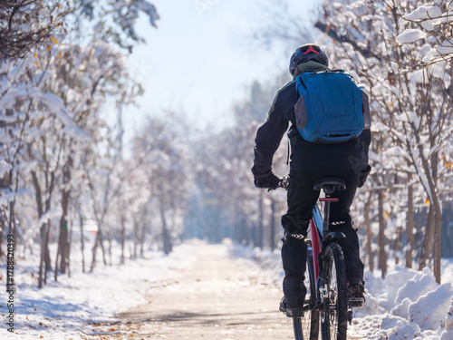 A rear view of a man riding a bicycle along a city bike path on a winter day. He is wearing a bicycle helmet. Active lifestyle in winter.