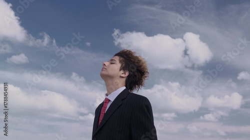 Portrait of a handsome teenager in a business suit against a backdrop of a hill and blue sky, his face upturned and his curls fluttering in the wind. The camera pans