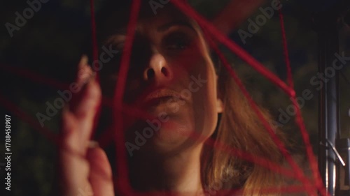 A woman looks at a weave of red threads in the dark and her face is illuminated by the flickering flame of a kerosene lamp