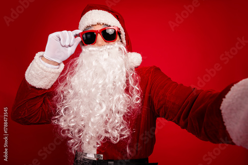 Fotografie Santa in red suit with white beard and red sunglass poses for a festive selfie a
