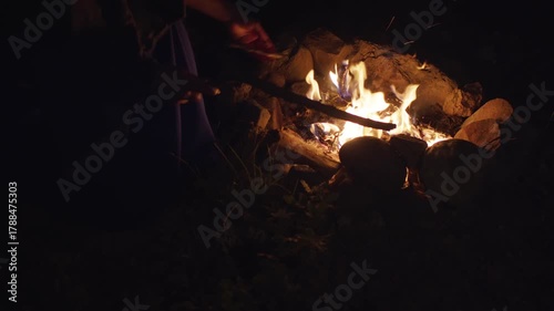 campfire scene scene, closeup of hands arranging firewood amidst rustic rocks under nighttime sky