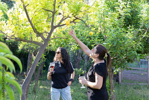 una mujer con lentes de sol con la mano alzada tocando un fruto de limón, atrás desenfocada 
otra mujer con lentes de sol sosteniendo una copa observando, rodeadas de naturaleza