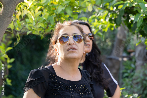 una mujer con lentes de sol en primer plano mirando hacia arriba, atrás desenfocada otra mujer con lentes de sol, rodeadas de naturaleza