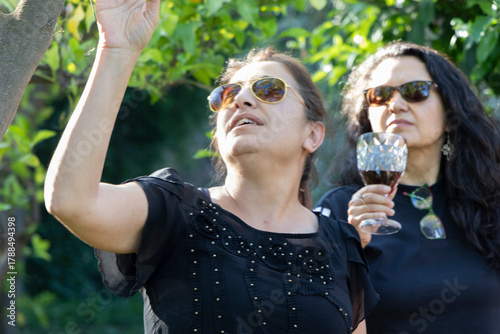 una mujer con lentes de sol en primer plano con la mano alzada y mirando hacia arriba, atrás desenfocada 
otra mujer con lentes de sol sosteniendo una copa observando, rodeadas de naturaleza