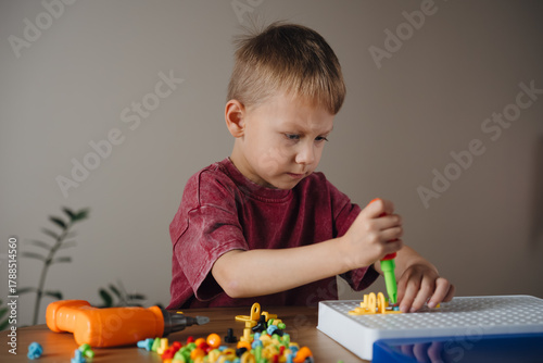 A young boy is carefully assembling a vibrant model using building blocks. He holds a green tool and shows concentration, working quietly at a wooden table in a bright room.