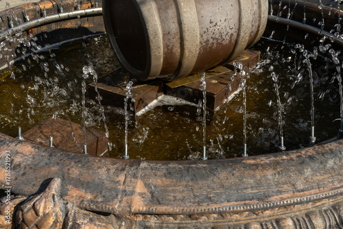 Water fountain with wooden barrel and circular jets in outdoor setting. Rustic barrel fountain with metal stand and water jets in public plaza with tables and chairs under natural daylight.