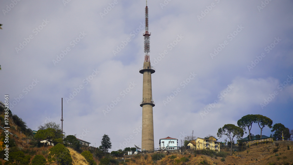 Fototapeta premium A tall, concrete communication tower with red & white sections stands on a hillside under a partially cloudy sky. Small,residential-style buildings are scattered around the base of the hill in kasauli