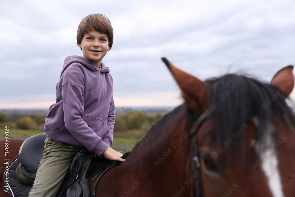Fototapeta premium Equine assisted therapy. Little boy riding beautiful horse outdoors. Lovely domesticated pet