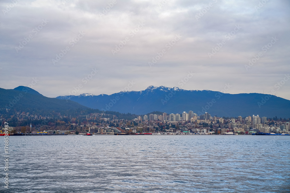 Fototapeta premium A panoramic view of a coastal city beneath expansive mountains, with shoreline homes, towers, and calm harbor water forming a balanced scenic landscape.