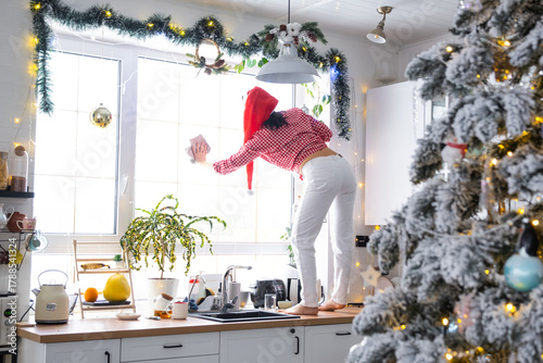 A woman in a Santa hat cleans a kitchen window with Christmas decorations, preparing for the New Year, decorating and tidying up the house